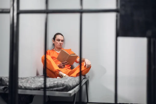 Um jovem recluso com uma cara tatuada está a ser treinado remotamente numa cela. Lê um manual sentado em um beliche. — Fotografia de Stock