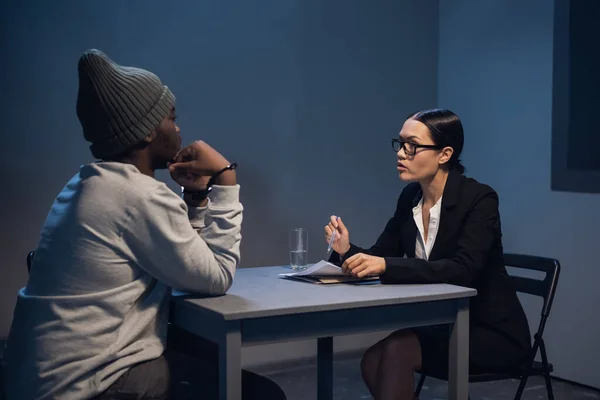 Uma jovem advogada consulta seu cliente na delegacia de polícia, um cara negro com um boné e algemas. — Fotografia de Stock