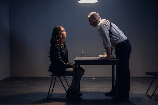 A white male police detective talks to a suspect in a drug trafficking case who is sitting in an interrogation room in handcuffs.