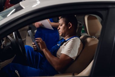 Tulum giymiş siyah bir adam arabada tablet kullanıyor.