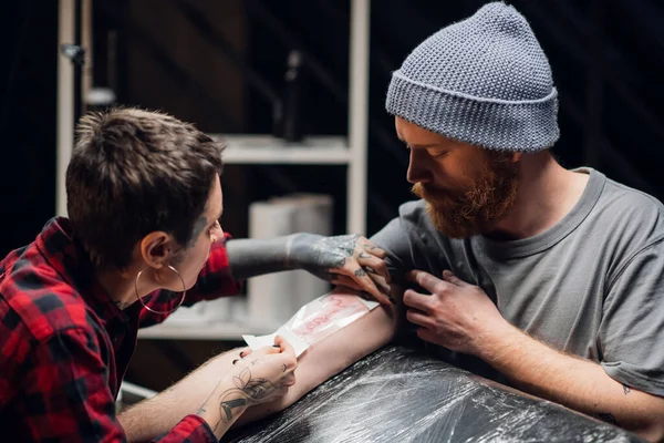 Joven artista informal del tatuaje de la muchacha pone un vendaje con vaselina en la mano de un hombre joven después de aplicar con éxito un tatuaje en su mano. — Foto de Stock