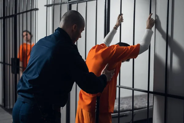 An armed guard searches a newly arrived criminal in a prison corridor against a backdrop of bars. — Stock Photo, Image