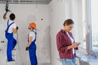 Tamirciler matkap ve merdiven kullanır, kadın telefonda yazar.