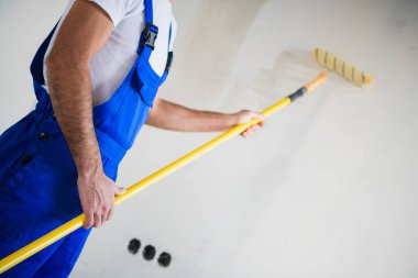 Yakından, tulumlu bir işçi duvarları beyaza boyuyor.