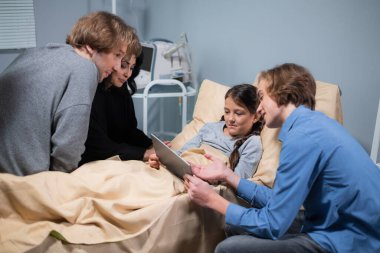 Yatağında dijital tabletle ve akrabalarıyla sağlık sorunları olan bir kız öğrenci.