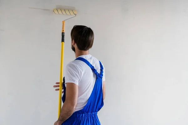 파란 작업복을 입은 노동자가 로울러 로 벽을 칠하고, 뒤에서 바라본다 — 스톡 사진
