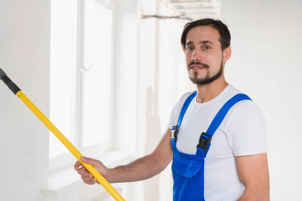 Hübscher Bauarbeiter in Overalls bemalt die Wand mit einer Walze — Stockfoto