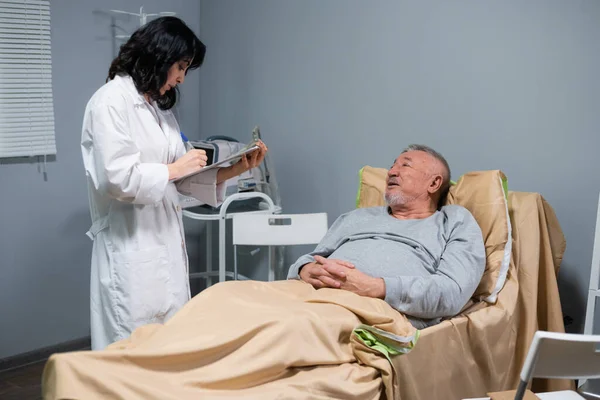 En læge holder en udklipsholder og tager noter, mens hun taler med sin patient på en hospitalsafdeling - Stock-foto
