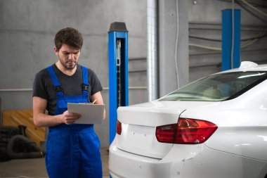 Elinde bir pano olan tamirci arabayı inceliyor.