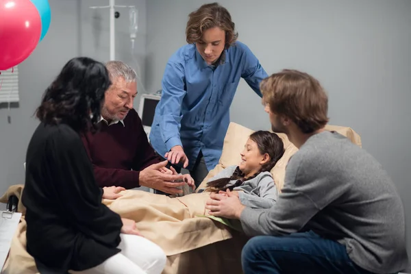 Una familia visitando a una niña bonita, que está acostada en la cama en un hospital —  Fotos de Stock