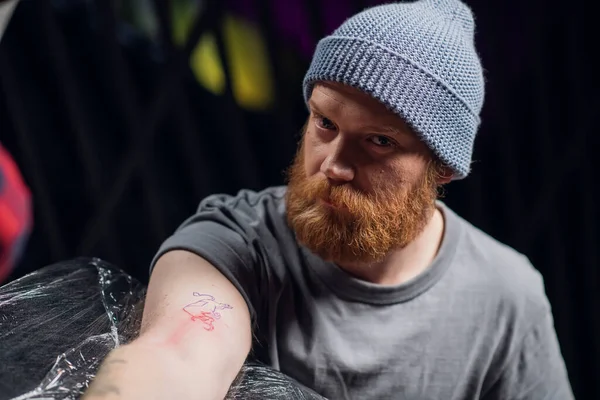 A young hipster in a knitted hat with a beard in a tattoo parlor in the process of getting his first bull tattoo