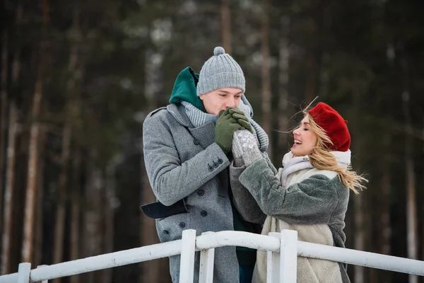 Um jovem aquece as mãos de sua namorada durante um passeio de inverno na ponte sobre um lago da floresta — Fotografia de Stock