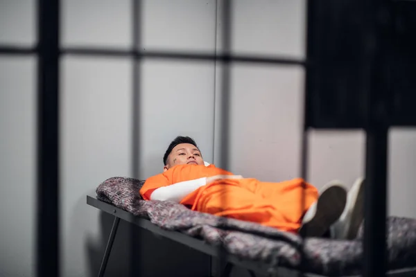 Een gevangene in een Aziatische gevangenis ligt op een bed in een rood gevangenenuniform in een eenzame cel.. — Stockfoto