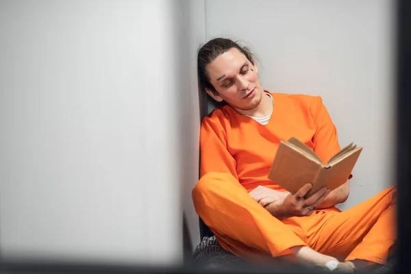 A dangerous mad criminal reads a book sitting on a bunk. In the foreground is the grid of a single cell. — Stock Photo, Image