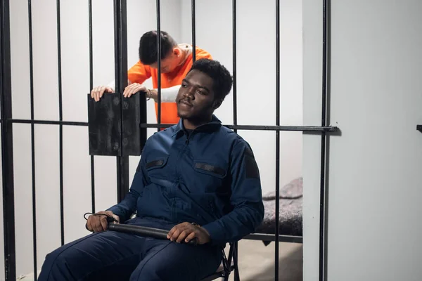 Un gardien de prison noir est assis devant des cages avec des criminels dangereux sur une chaise avec un bâton. — Photo