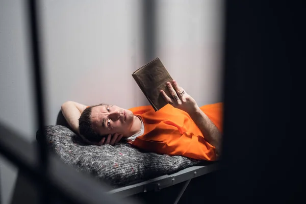 A dangerous serial killer with tattoos on his face in a cell reads the Holy Bible lying on a bunk. — Stock Photo, Image