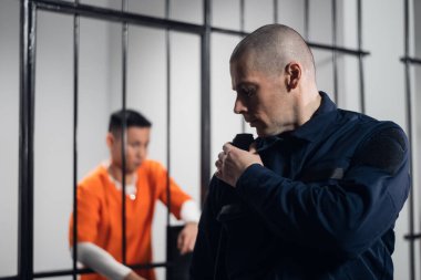 Tam teçhizatlı bir gardiyan telsizdeki ortağıyla iletişim kuruyor. Arka planda, hücrede bir mahkum