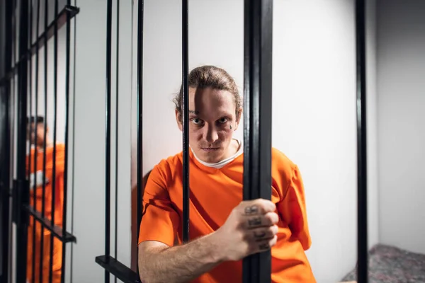 Un maníaco criminal desesperado con tatuajes en una celda solitaria se mantiene tras las rejas. Una mirada dura a la cámara. —  Fotos de Stock
