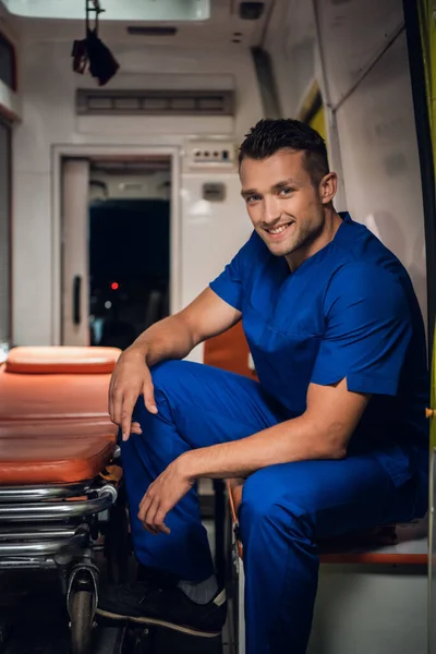 Um retrato de um paramédico sorrindo em um uniforme sentado na parte de trás de uma ambulância com uma maca — Fotografia de Stock