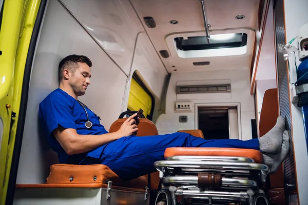 Мужчина-парамедик в синей форме отдыхает и разговаривает с кем-то по видеосвязи со своего смартфона в машине скорой помощи — стоковое фото