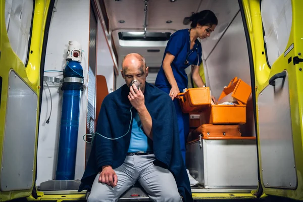 Un hombre herido sentado con una máscara de oxígeno en un coche ambulancia, una enfermera revisando su botiquín en el fondo — Foto de Stock