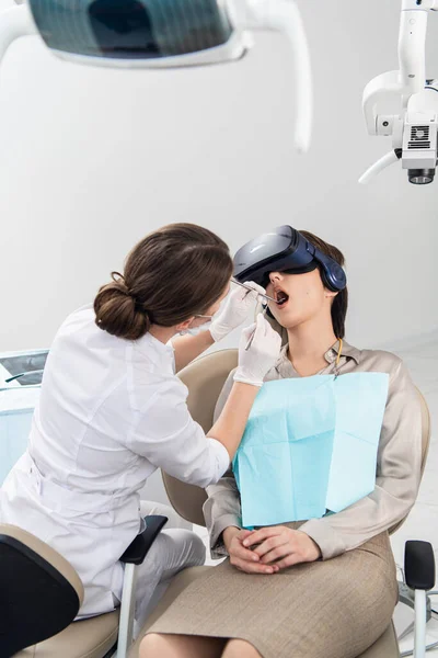 Dentystka wykonuje swoją pracę, podczas gdy pacjentka leży z otwartymi ustami, w okularach VR — Zdjęcie stockowe