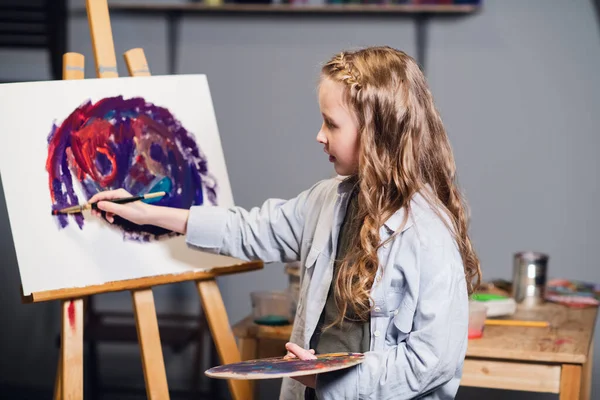 Una joven artista linda está aprendiendo a dibujar en un taller de arte. Se basa en un caballete con pinturas al óleo y un pincel. — Foto de Stock