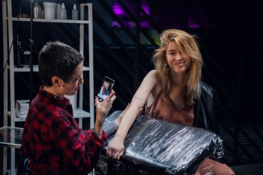 Dövme salonunda, bir dövme sanatçısı akıllı telefondaki çalışmalarının sonucunu fotoğraflıyor.