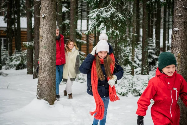 Девушка и мальчик в зимней одежде бегут в зимнем снежном лесу, их родители за ними — стоковое фото
