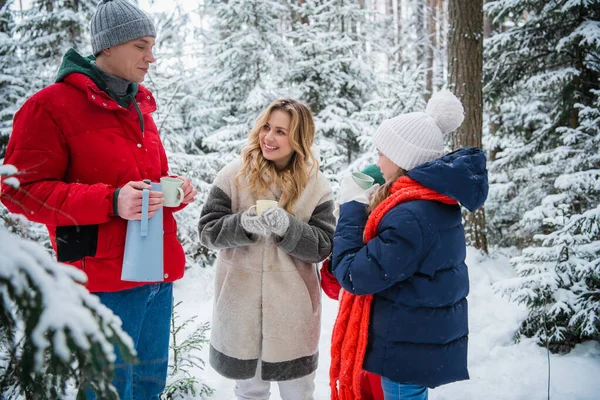 A healthy friendly family actively spends a weekend in the nature in a winter spruce forest during a beautiful snowfall and enjoys hot tea from hiking mugs. — стоковое фото