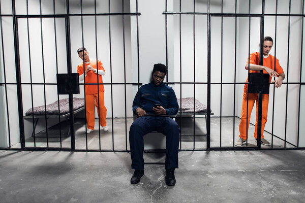 A strong black prison guard guards the cells with prisoners in the prison corridor