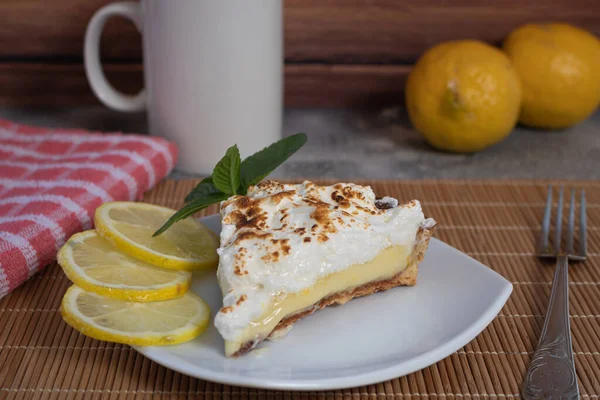 A slice of homemade lemon meringue pie on a plate, with lemons, napkin and cup of coffee completing the scene in a rustic setting and wooden planks background