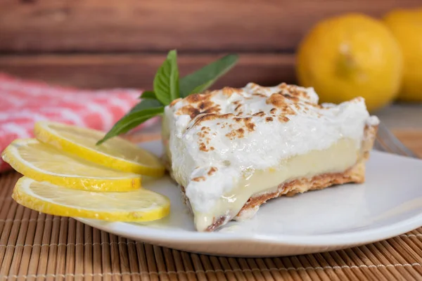 A slice of homemade lemon meringue pie on a plate, with lemons, napkin completing the scene in a rustic setting and wooden planks background