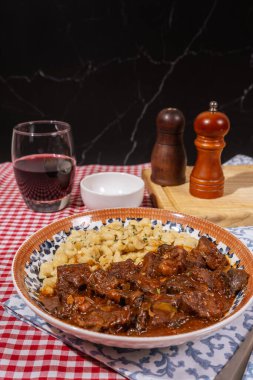 Buharda pişmiş gulaş, yanında bir bardak kırmızı şarap ile spaetzle köftesi ile servis edilir, rahatlatıcı ve lezzetli bir yemek deneyimi yaratır.