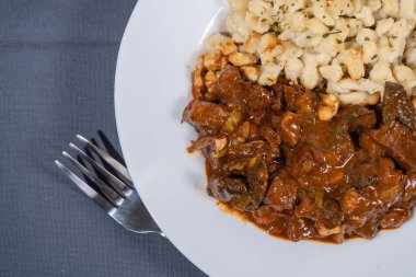 Macar tas kebabı, gri masa örtüsünün üzerinde duran çatalla birlikte beyaz bir tabakta spaetzle mantı ile servis edilir. Bu geleneksel yemeğin en üst görüntüsünü sunar.