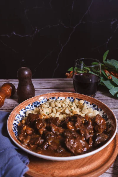Lezzetli Macar tas kebabı bir bardak kırmızı şarapla birlikte spaetzle köftesi ile servis edilir ve geleneksel bir aşçılık deneyimi yaratır.