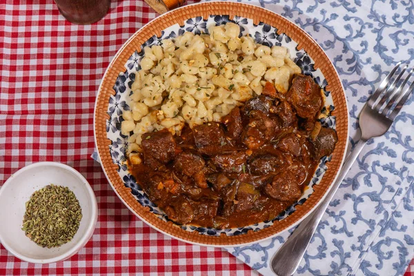 Macar tas kebabı, spaetzle ve baharatla dekoratif bir tabakta servis edilir, geleneksel mutfağı somutlaştırır.
