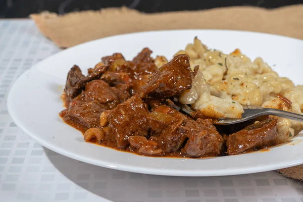 Beyaz bir tabakta sığır kebabı ve spaetzle yakın plan, bir parça spaetzle kaldıran bir çatalla.