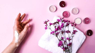 Nemlendirici krem süren bir kadın. Pembe arka planda küçük pembe çiçekli beyaz pamuk havlu ve aroma mumları, kopya alanı, cilt ürünleri, spa tedavisi için doğal kozmetik ürünleri.