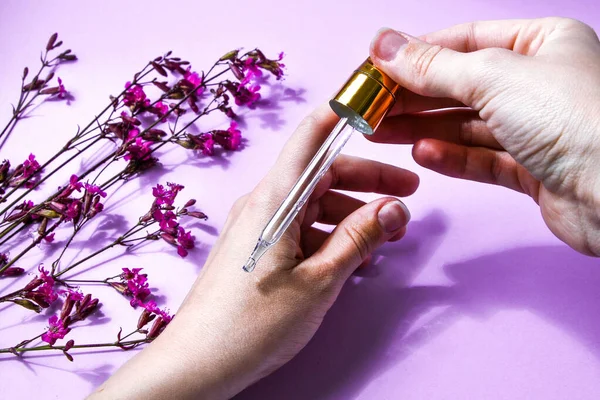 Woman dripping rose essential oil on hand against blurred flowers on pink background, closeup, white spa towel, beauty treatment, hand skin care, natural medicine, Women\'s hands applying oil with a drip