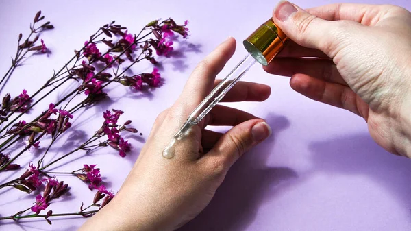 Woman dripping rose essential oil on hand against blurred flowers on pink background, closeup, white spa towel, beauty treatment, hand skin care, natural medicine, Women\'s hands applying oil with a drip