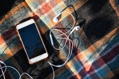 Smartphone with headphones on the cozy sweaters. Top view. Autumn flatlay. Outdoor listening podcasts. Sunlight clipart
