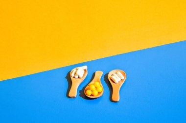 Ahşap kaşıkta haplar mavi ve sarı arka planda. Sert ışık ve gölgeler. Modern izometrik yaratıcı minimalizm kavramı. Vitaminler, prebiyotikler, probiyotikler. Sonbahar vitamin dozu.