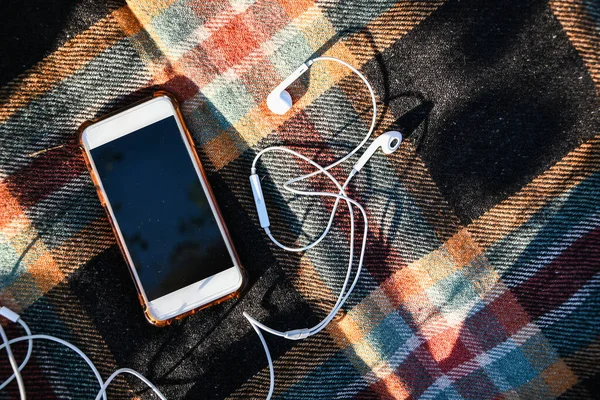 Rahat kazaklarında kulaklık olan akıllı bir telefon. Üst Manzara. Sonbahar düzlüğü. Açık hava dinleme podcastları. Güneş ışığı