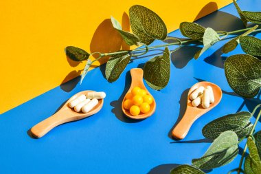 Ahşap kaşıkta haplar mavi ve sarı arka planda. Sert ışık ve gölgeler. Modern izometrik yaratıcı minimalizm kavramı. Vitaminler, prebiyotikler, probiyotikler. Sonbahar vitamini dozu. Okaliptüs