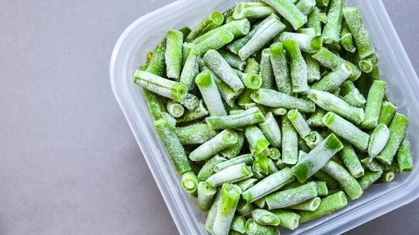 Frozen green beans in a plastic container for long-term storage. Deep freezing of vegetables. Frozen food vegetables on grey background. Copy space for text. Stocking up vegetables for winter storage