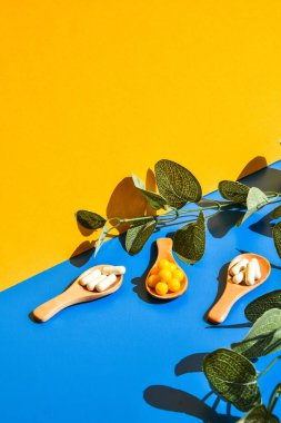 Ahşap kaşıkta haplar mavi ve sarı arka planda. Sert ışık ve gölgeler. Modern izometrik yaratıcı minimalizm kavramı. Vitaminler, prebiyotikler, probiyotikler. Sonbahar vitamini dozu. Okaliptüs