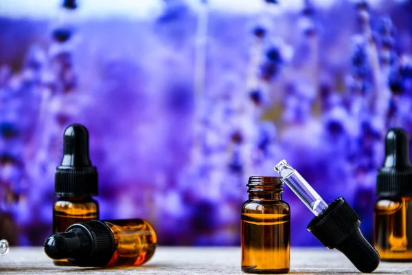 Natural essential oil dripping from pipette into bottle against lavender flowers, closeup. Bottles with natural lavender essential oil on wooden table against blurred background