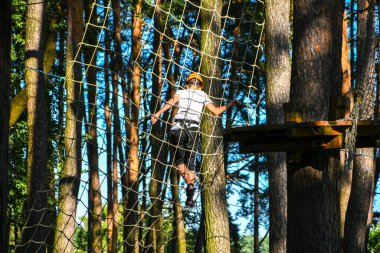 Yüksek tel parka tırmanma macerası. Yakın plan. Miğferli genç çocuk eğleniyor ve macera parkında oynuyor, ipleri tutuyor ve tahta merdivenlere tırmanıyor. aktif yaşam tarzı konsepti. Yaz tatili aktiviteleri
