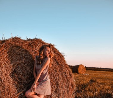 Kırsal kesimde, sabah güneşinde samanlıkta kızıl saçlı seksi bir kadının portresi. Elbise giymiş güzel bir kadın samanlıkta oturuyor. Cottagecore 'da. Saman yığınlarıyla yaz tarlasında dinlenmek. Temiz hava kırsal kesimde. pastoral yaşam